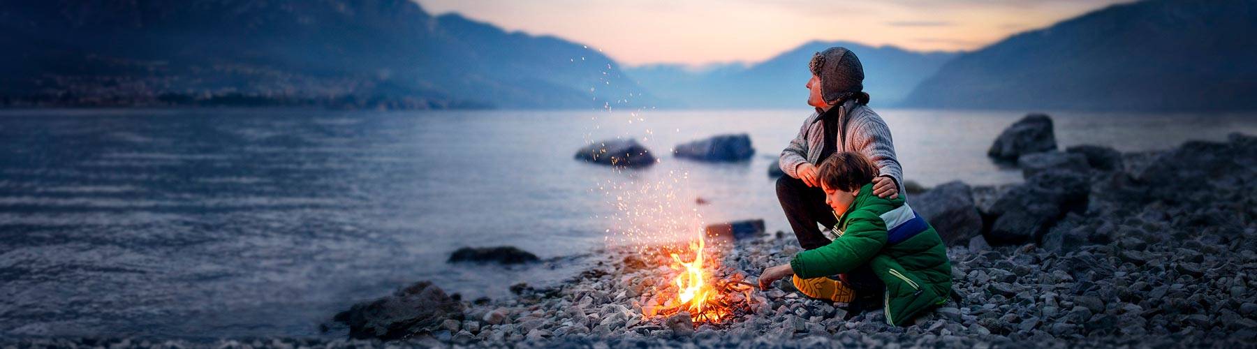 Imagen paisajística de dos personas disfrutando de un fuego junto al lago al atardecer